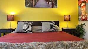Una cama con un edredón rojo, dos almohadas a rayas blanco y negro y un plaid de piel. Hay dos mesitas de noche con lámparas y un cuadro con una escena de playa sobre la cama.