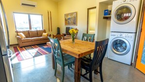 A compact living space with a sofa, dining table, chairs, a washing machine, and a painting on the wall.