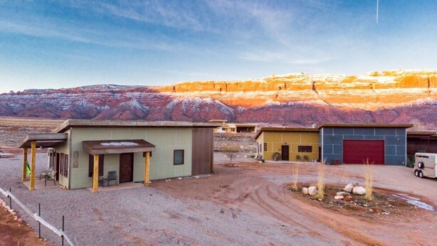 A small building with a red garage door, a dirt driveway, and a mountainous backdrop.