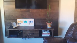 A living room with a mounted TV, wooden shelves, a framed artwork, a small table, a potted plant, and a leather sofa.