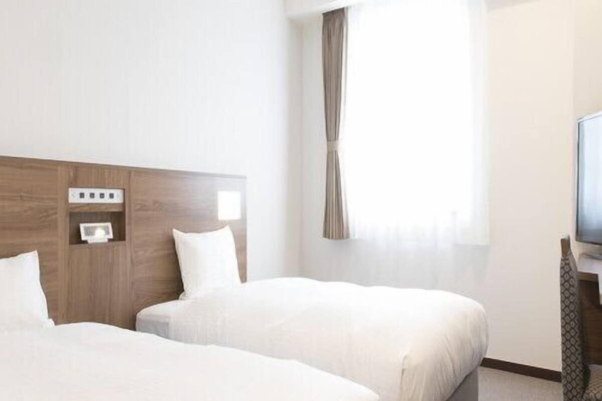 A hotel room with two beds, a television, a power outlet, and a window with curtains.