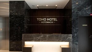 Uma área de recepção com balcão de mármore, um letreiro com a inscrição "TOHO HOTEL MOTOMACHI" e uma porta com a inscrição "ESCRITÓRIO" à direita.