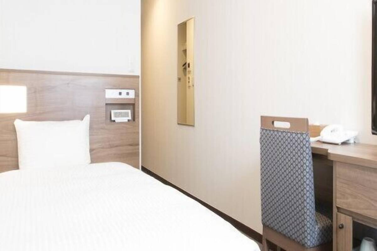 A hotel room with a bed, a desk with a telephone, a mirror, and a wall-mounted heater.