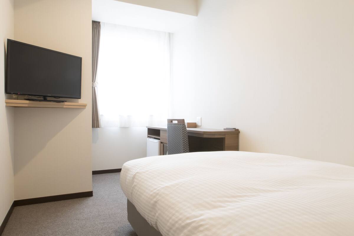 A hotel room with a bed, a flat-screen TV mounted on the wall, a desk with a chair, and a window with curtains.