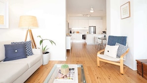 Un salon moderne avec un canapé, un fauteuil et une table basse. On aperçoit une cuisine au fond.