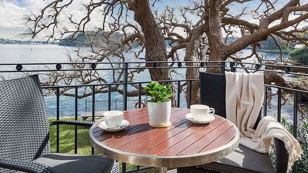 Une table ronde en bois, sur laquelle sont posées deux tasses blanches et une plante en pot, est placée sur un balcon d’où l’on peut admirer un lac et des arbres.