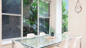Un espace repas moderne avec une table en verre et des chaises blanches, de grandes fenêtres donnant sur un paysage tropical et une horloge au mur.