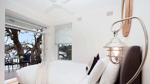 Une chambre à coucher avec un lit, un ventilateur de plafond, une fenêtre avec des stores et une vue sur des arbres et une étendue d’eau.