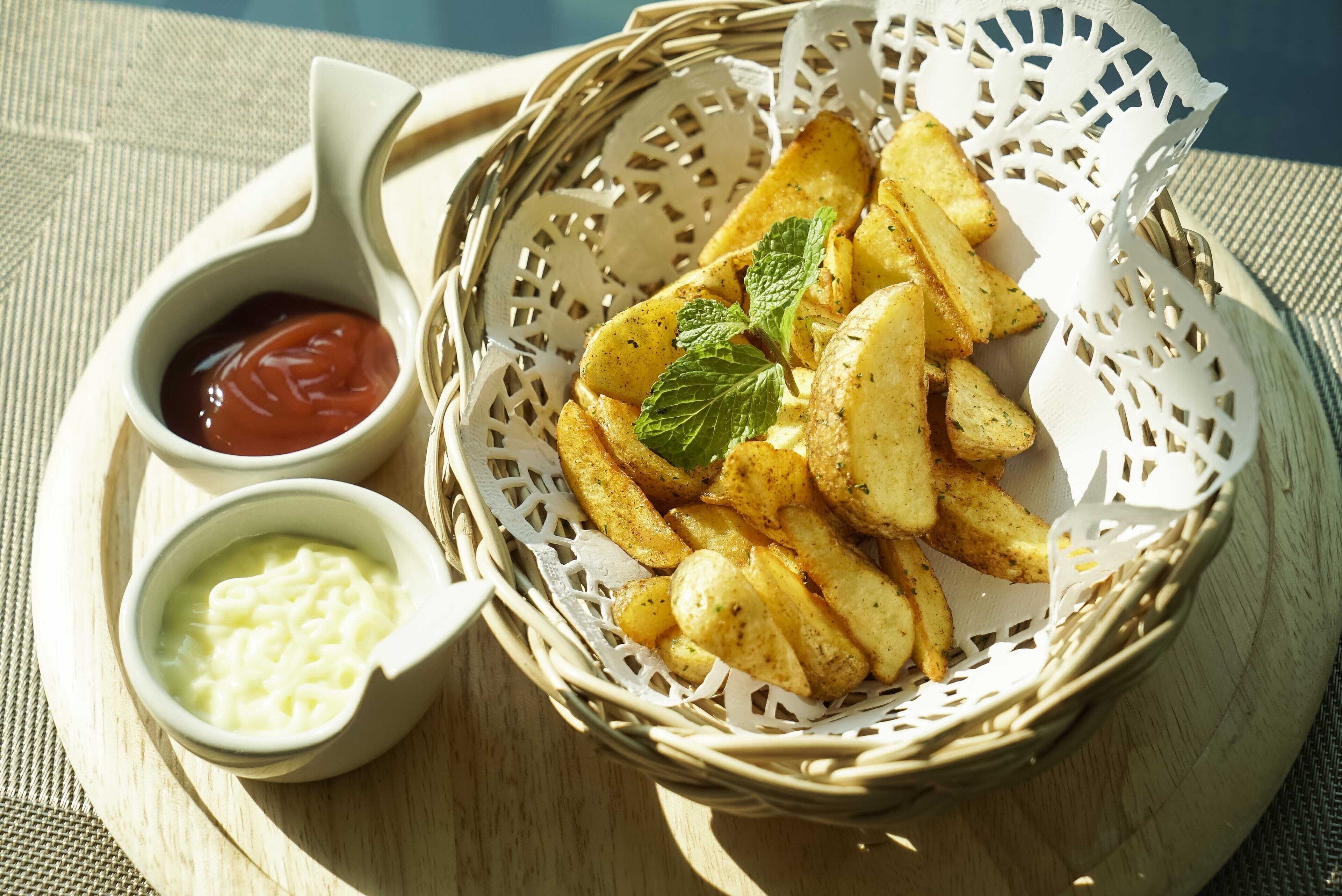 A basket of seasoned potato wedges with two dipping sauces, one red and one white, on a woven mat.