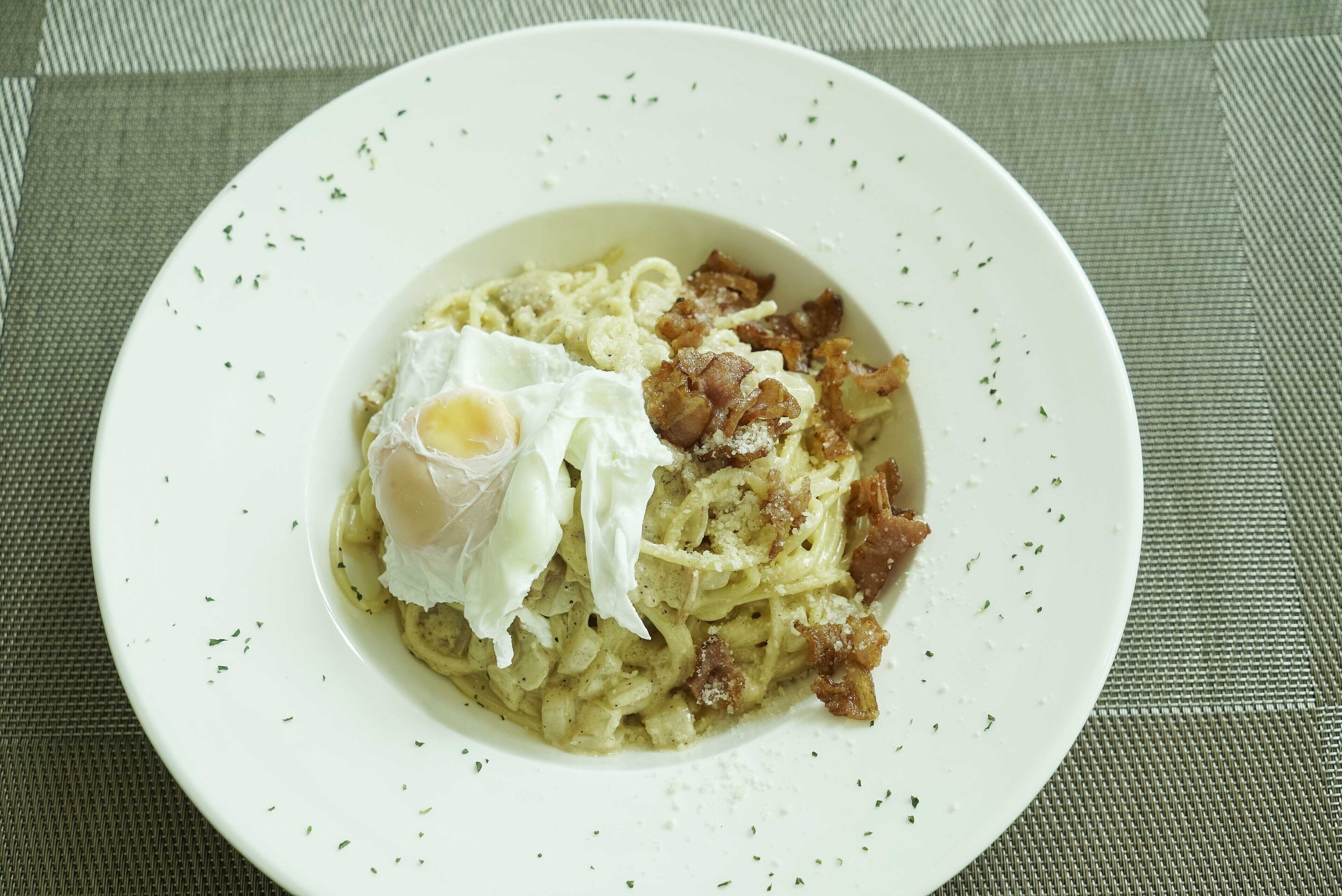 Un plato de comida con pasta, tocino y un huevo pochado sobre un plato blanco.