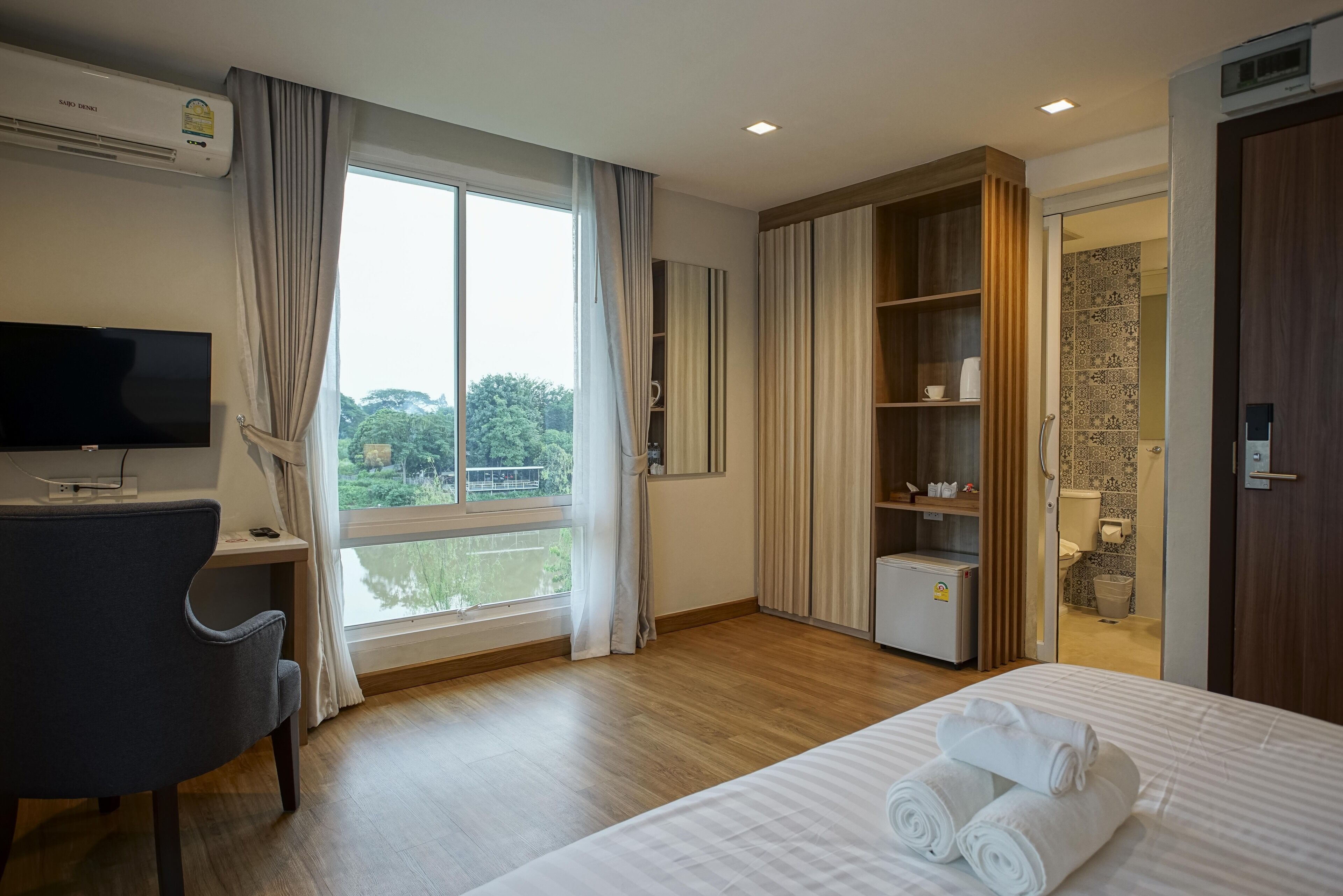 A hotel room with a bed, a desk with a computer, a TV, a window with curtains, a wardrobe, and a bathroom visible through an open door.