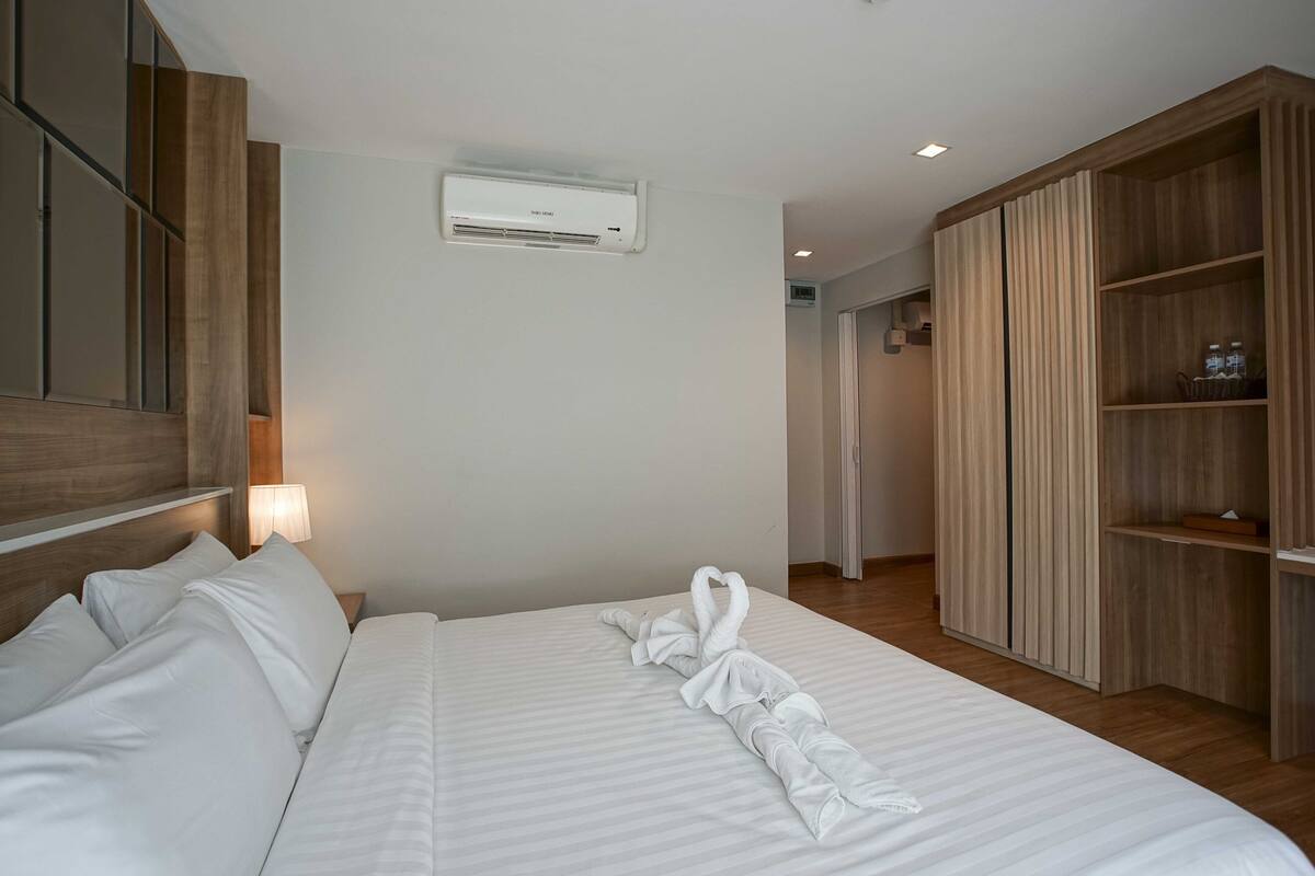 A hotel room with a neatly made bed, a wall-mounted air conditioner, a wooden wardrobe, and a bedside lamp.
