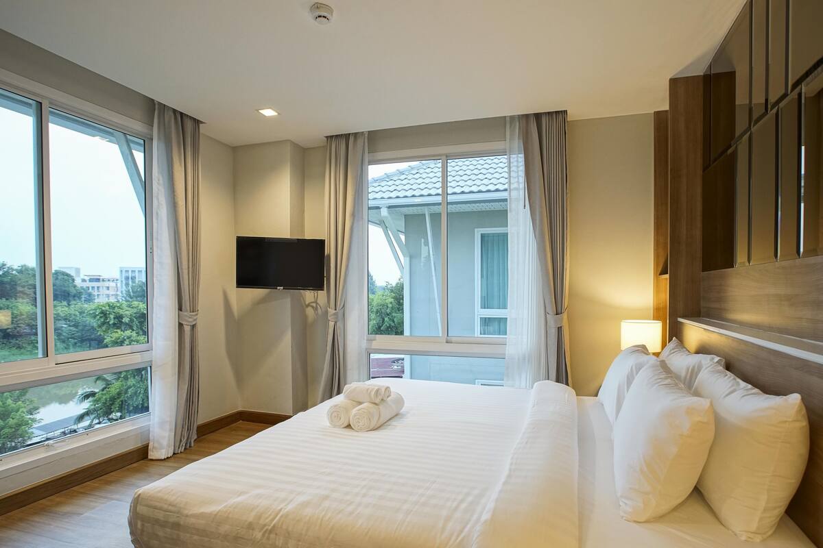 A hotel room with a large bed, a flat-screen TV mounted on the wall, and a view of the cityscape through large windows.