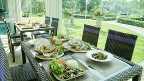 A dining table set with various dishes, surrounded by wicker chairs, in a well-maintained garden with a fountain and statues. - Nantra Chiangmai Riverfront Hotel (Chiang Mai)