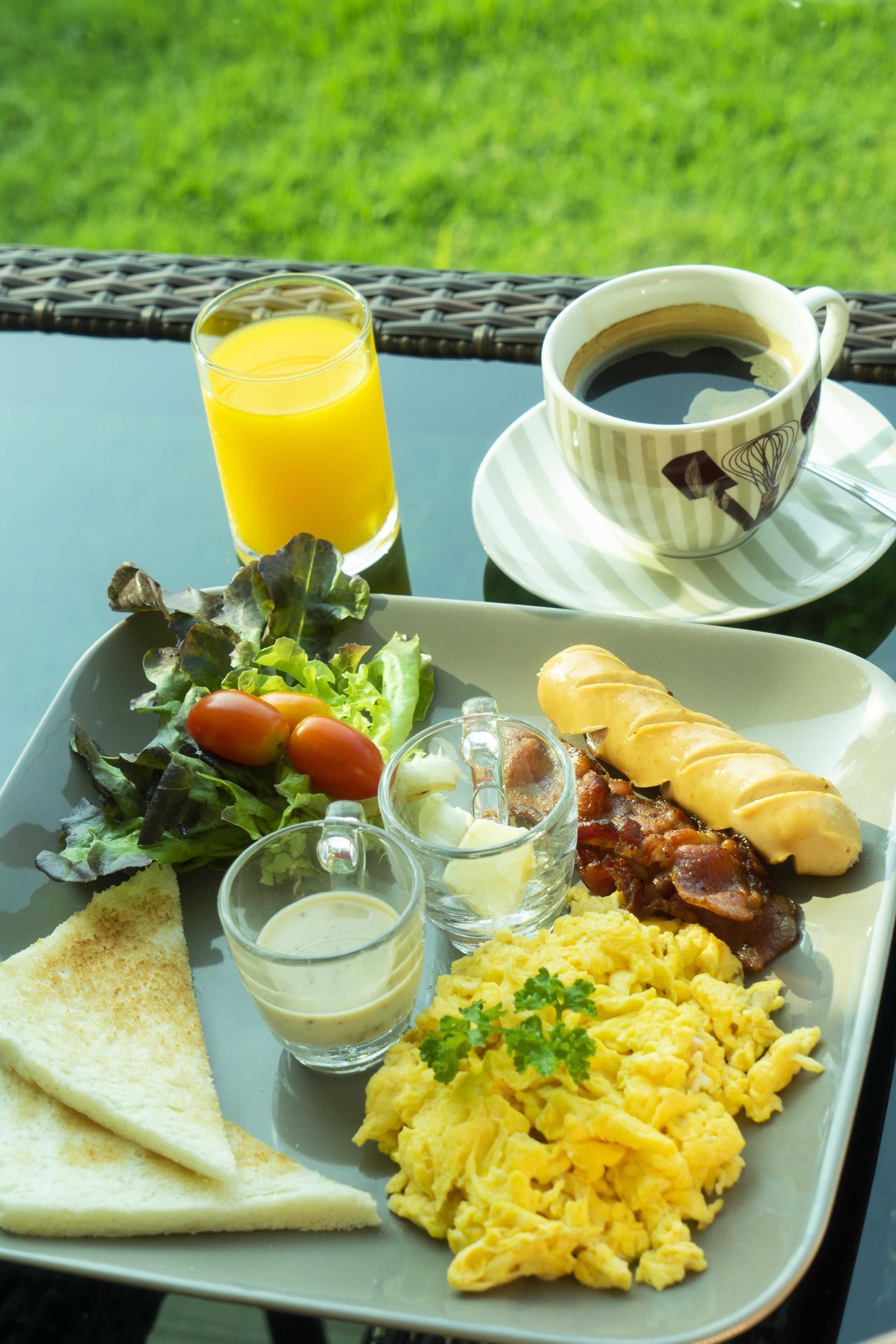 Un vassoio per la colazione con uova strapazzate, pancetta, pane tostato, un'insalata e una tazza di caffè su un tavolo, con un prato verde sullo sfondo.