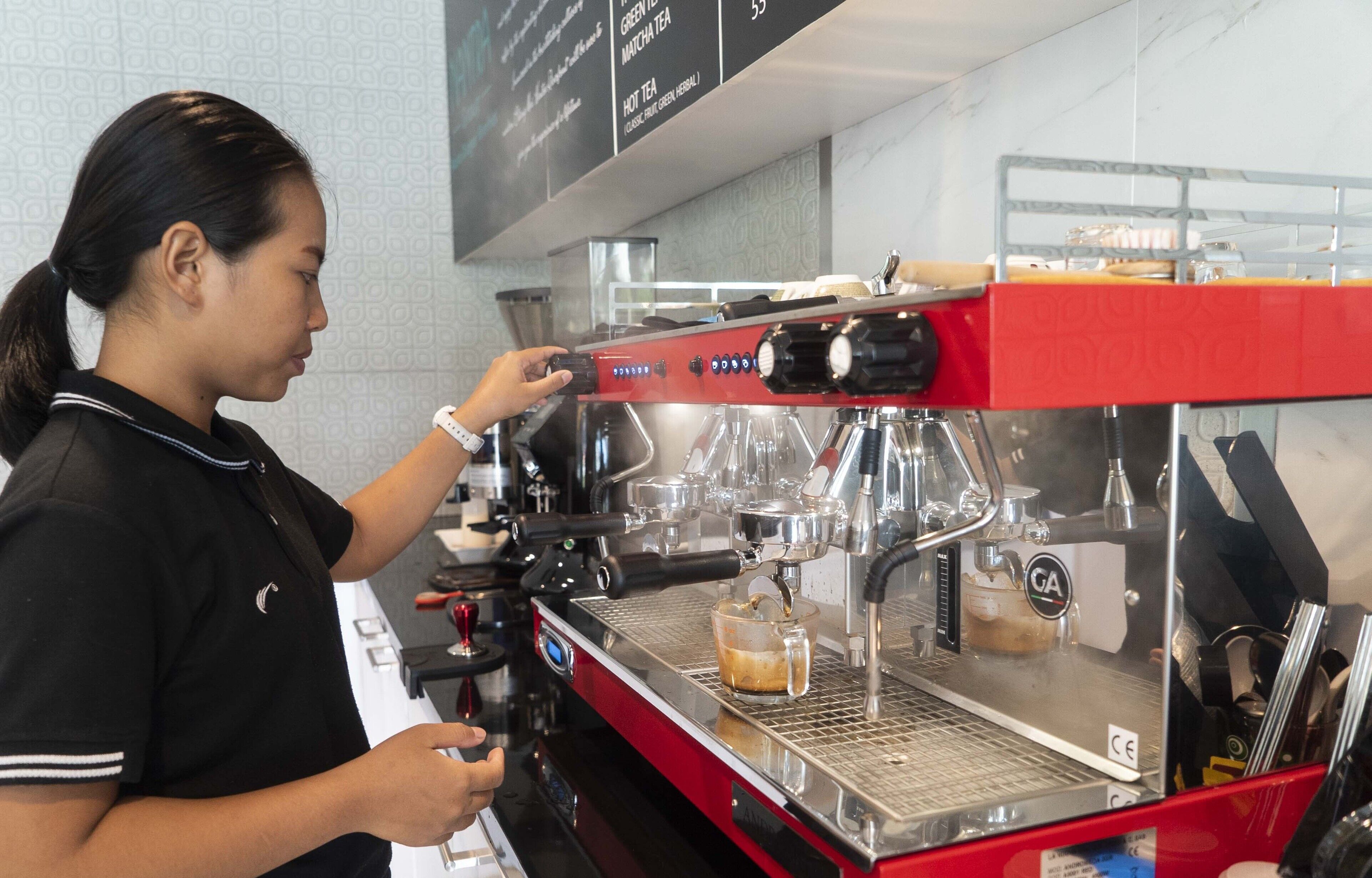 Una persona operando una máquina de café espresso roja con pantalla digital, una estación para espumar leche y una taza de vidrio sobre la encimera.