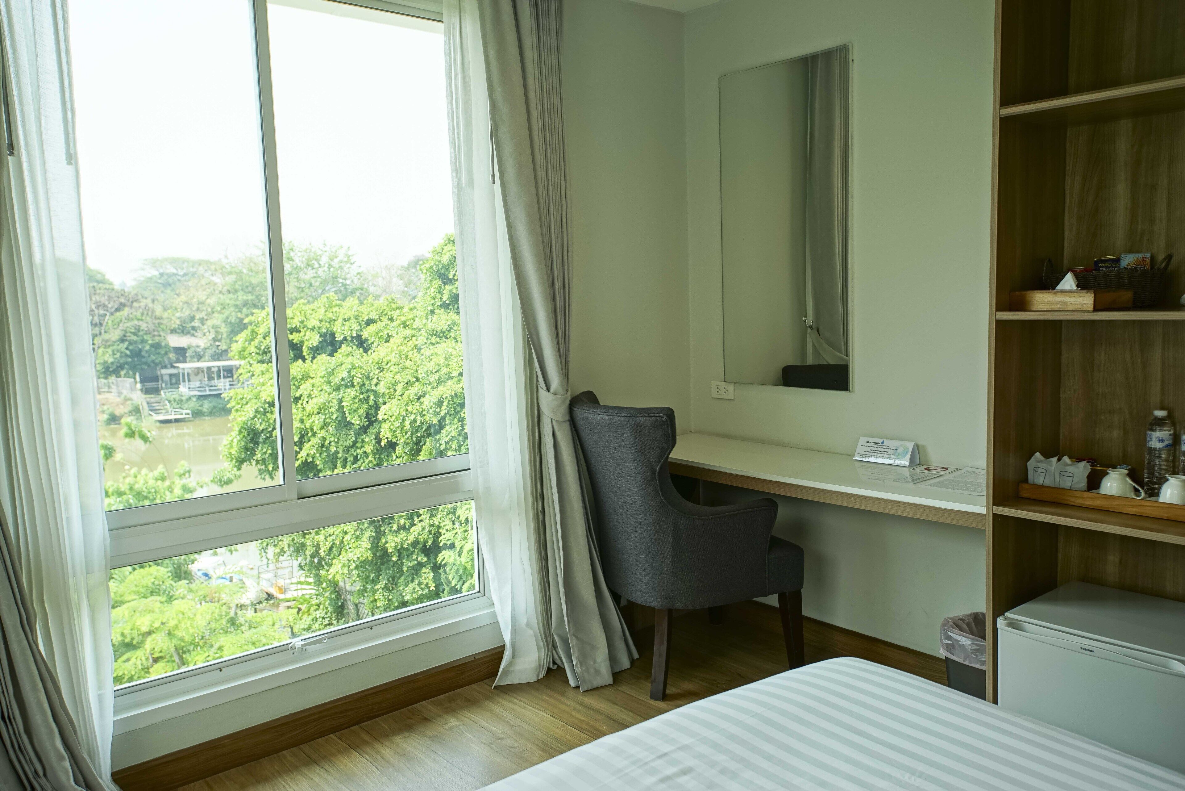 A hotel room with a large window, a desk, a chair, a bed, a mirror, and a small refrigerator.