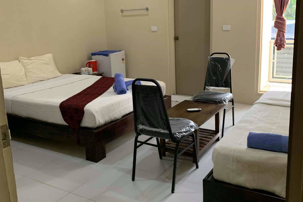 A hotel room with two beds, a table, a chair, an air conditioner, and a window with curtains.