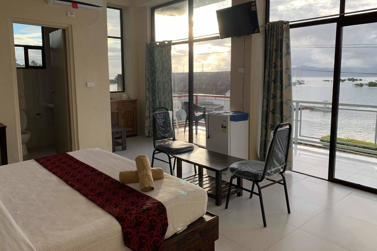 A hotel room with a bed, a desk, a chair, a TV, and a view of the sea.