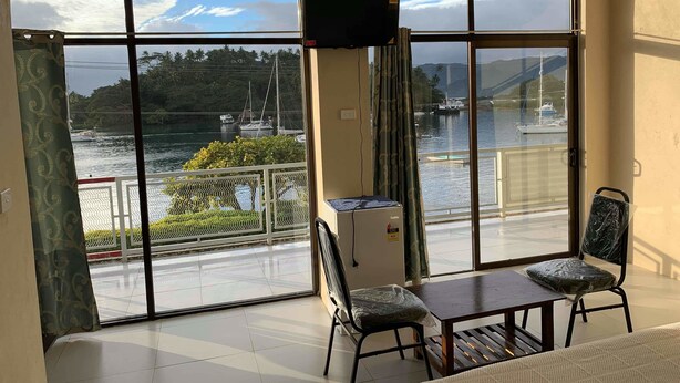 A room with large windows overlooking a marina, a television, and a dining area with chairs and a table. - Bay View Barbers Lodge (Savusavu)
