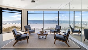 Quarto moderno com uma janela grande com vista para uma praia, mobiliado com cadeiras e uma mesa de centro.