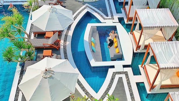 Zona de piscina con múltiples sillas de descanso y sombrillas, rodeada de exuberante vegetación y una piscina central.