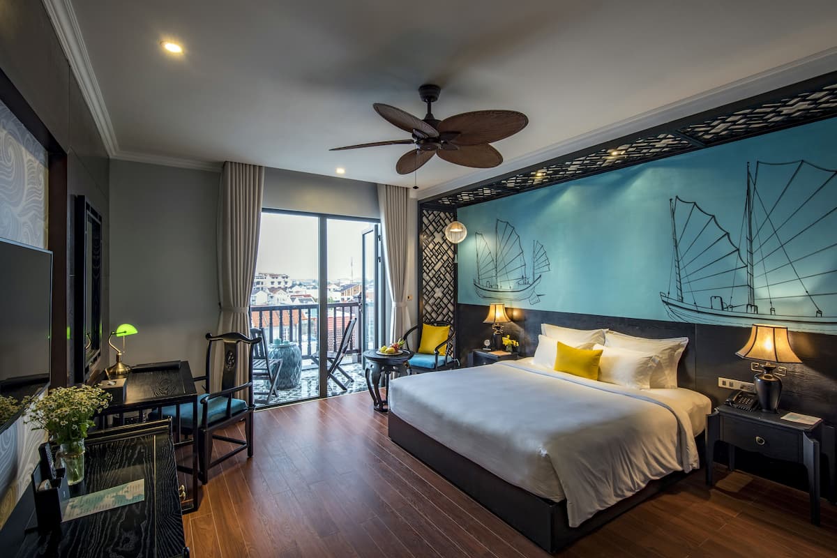 Ein Hotelzimmer mit einem großen Bett, einem Deckenventilator, einem Balkon mit Aussicht und einem Wandbild mit Segelbooten.
