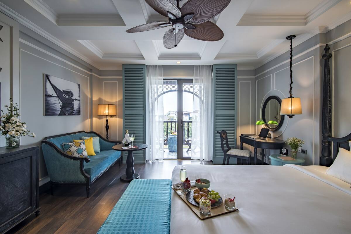 Ein Hotelzimmer mit Bett, Sofa, Schreibtisch, Stuhl, Deckenventilator und Blick ins Freie.
