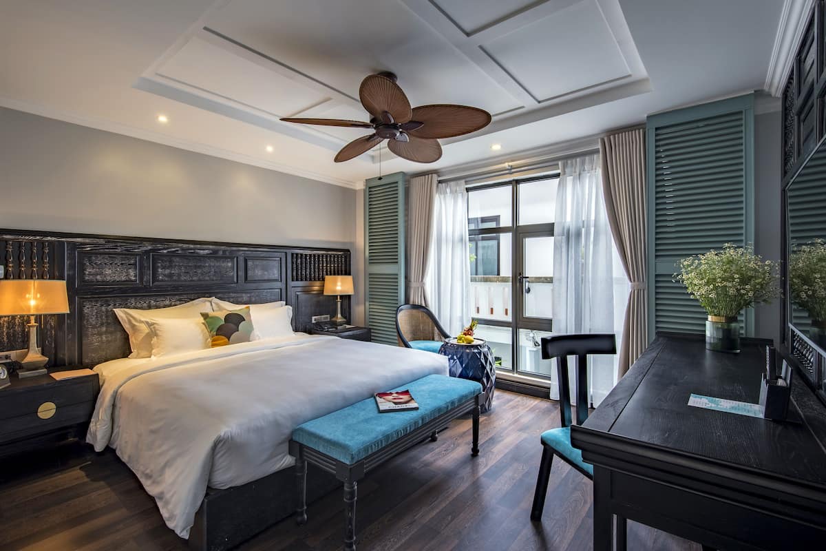 Ein Hotelzimmer mit einem großen Bett, einem Schreibtisch, einem Sessel, einem Deckenventilator und einem Vorhangfenster.