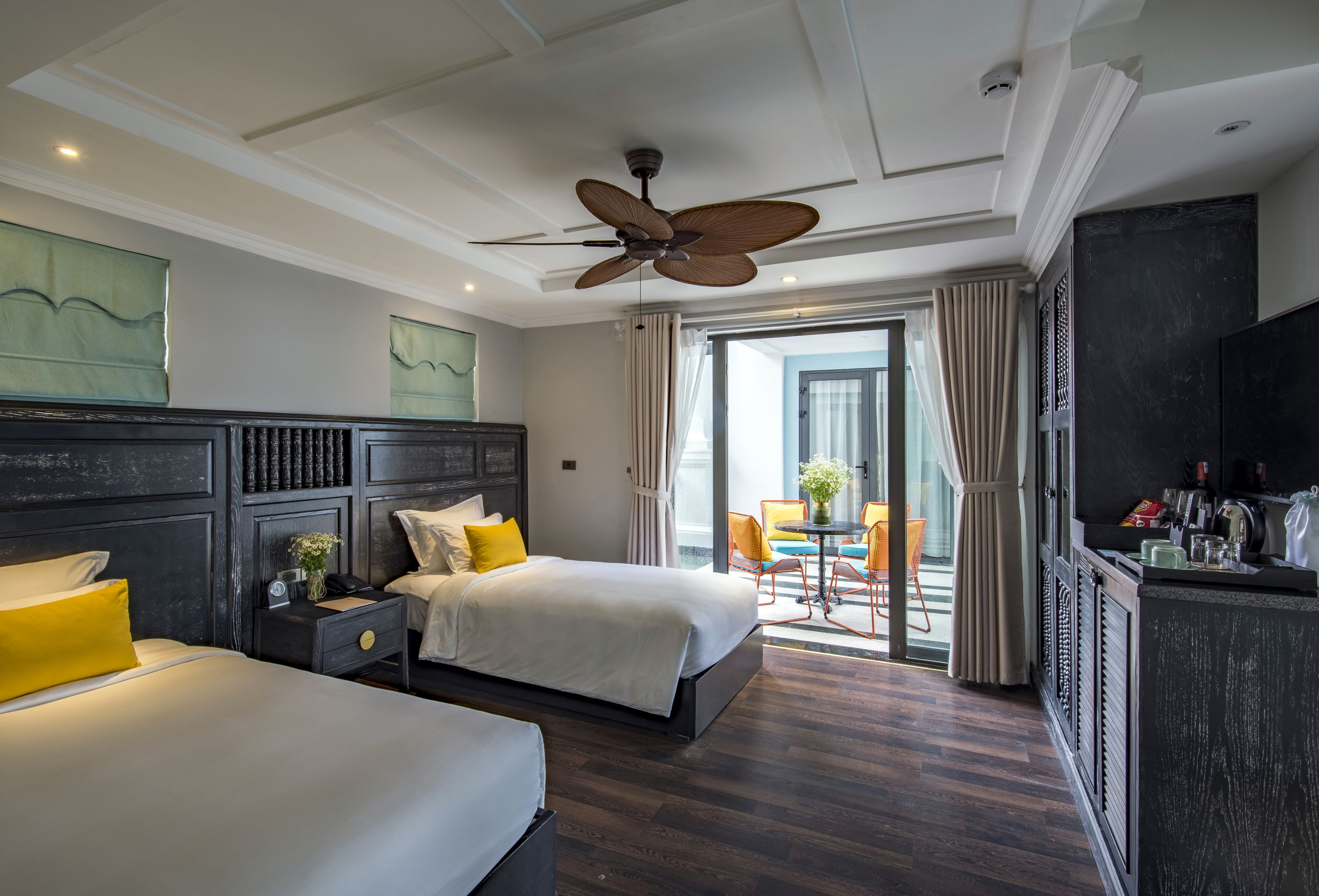 Quarto de hotel com duas camas, ventoinha no teto, varanda com cadeiras e minibar.