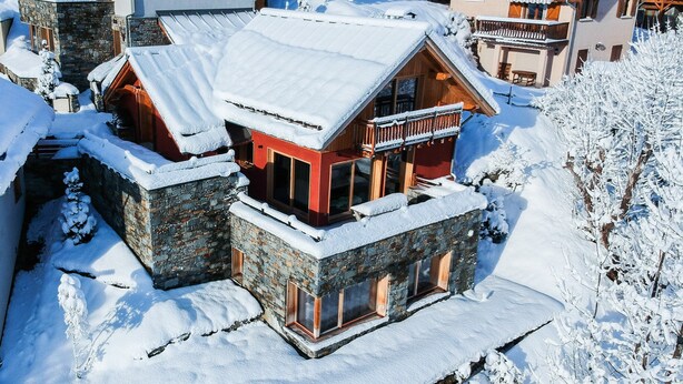 En snötäckt stuga med en fasad av sten, en träbalkong och en fri utsikt över det snöklädda landskapet.