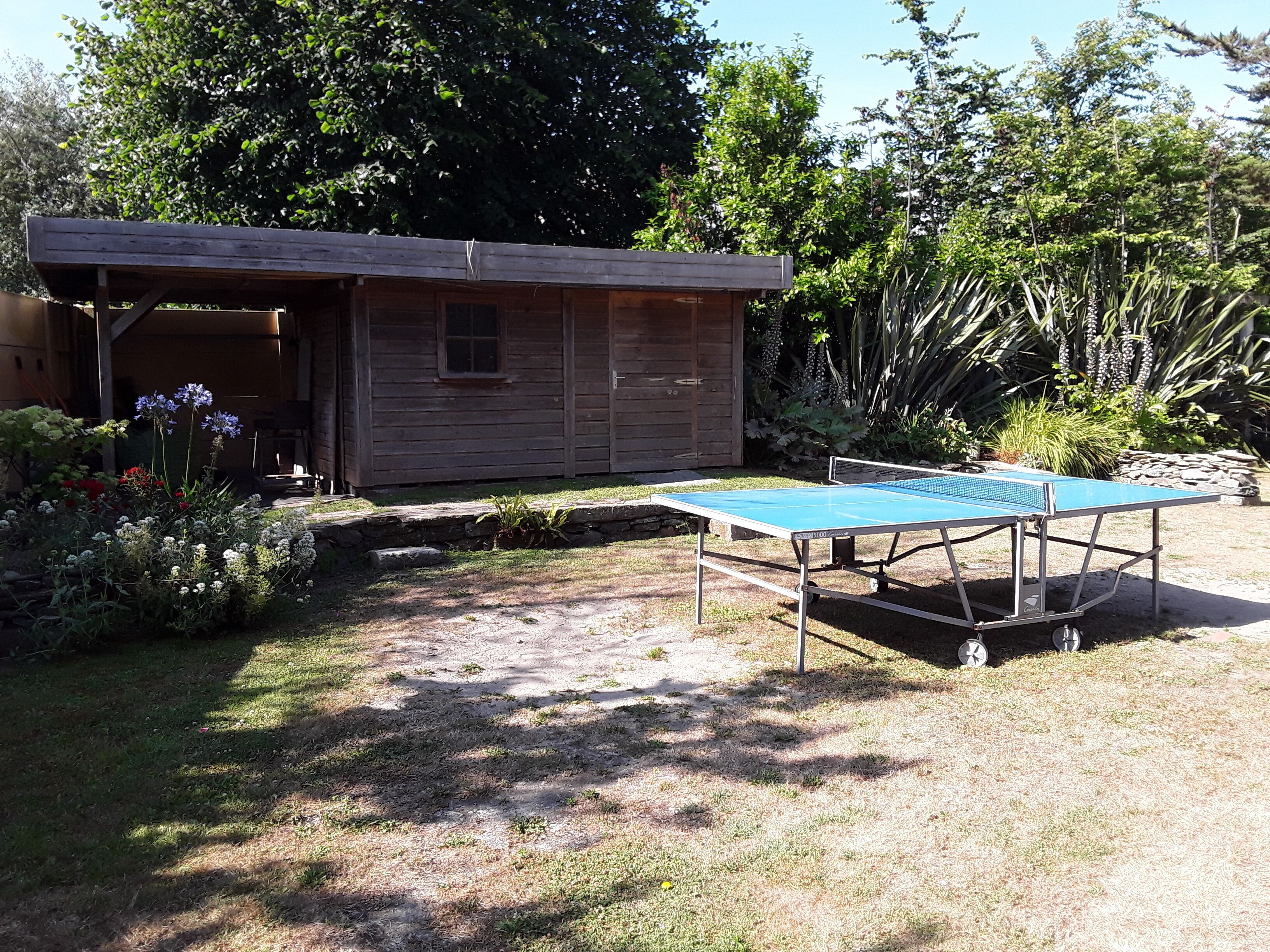 Un bâtiment en bois doté d’un porche couvert, d’une table de ping-pong et d’un jardin où poussent diverses plantes et fleurs.