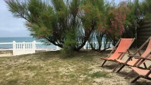 A beachside area with a row of orange lounge chairs, a white fence, and a tree with green foliage and pink flowers. - House ALL COMFORT, NICE GARDEN, FEET IN THE WATER, DIRECT BEACH ACCESS (La Hague)