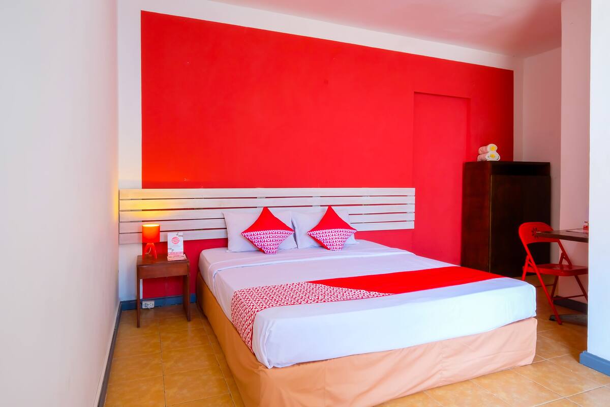 A hotel room with a red accent wall, a bed with white and red bedding, a nightstand with a lamp, and a red chair.