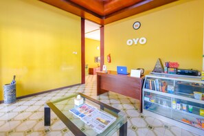 A reception area with a yellow wall, a reception desk, a glass display case with items, and a coffee table with a newspaper. - OYO 1206 Lombok Guest House (Mataram)