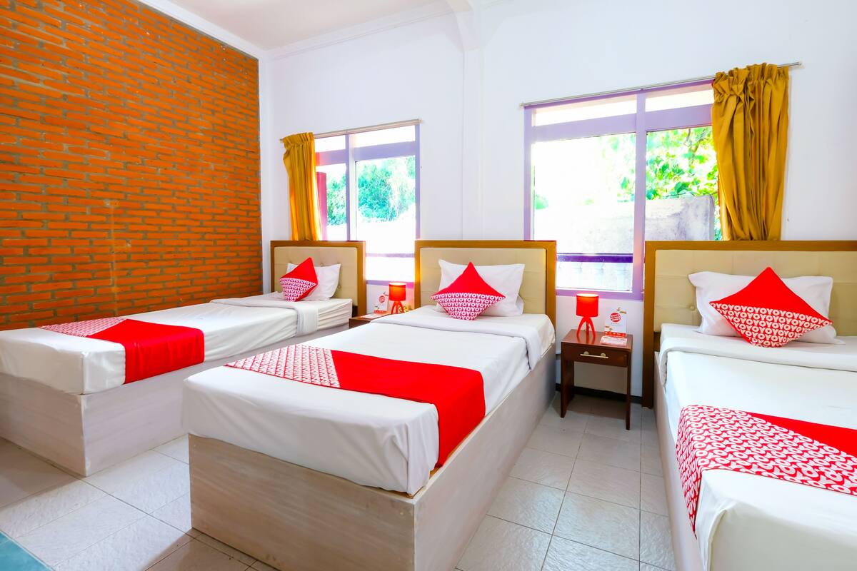 A hotel room with four single beds, each with white bedding and red accents, a brick wall, and large windows with yellow curtains.
