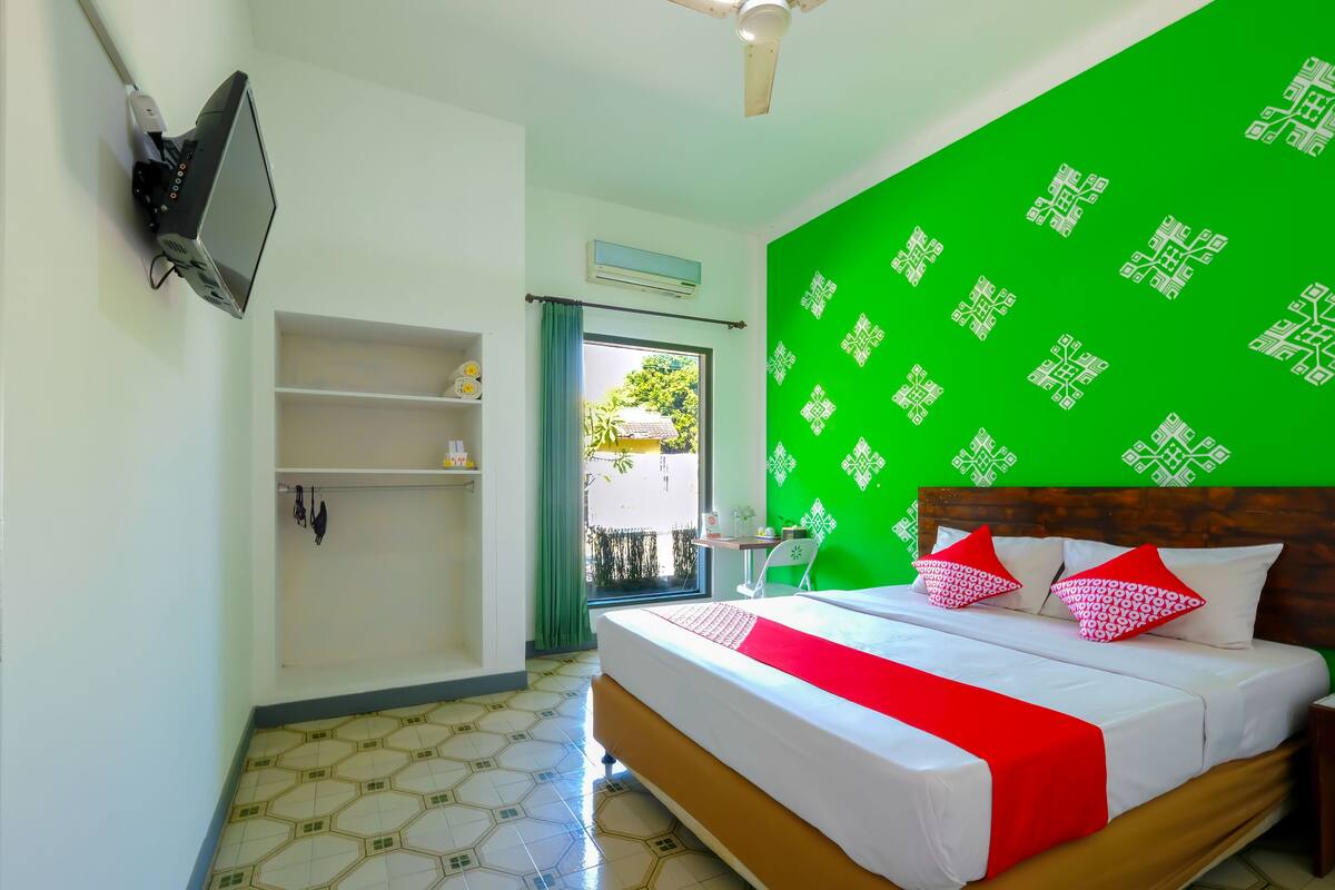 A hotel room with a bed, a TV mounted on the wall, a green wall with a geometric pattern, and a tiled floor.
