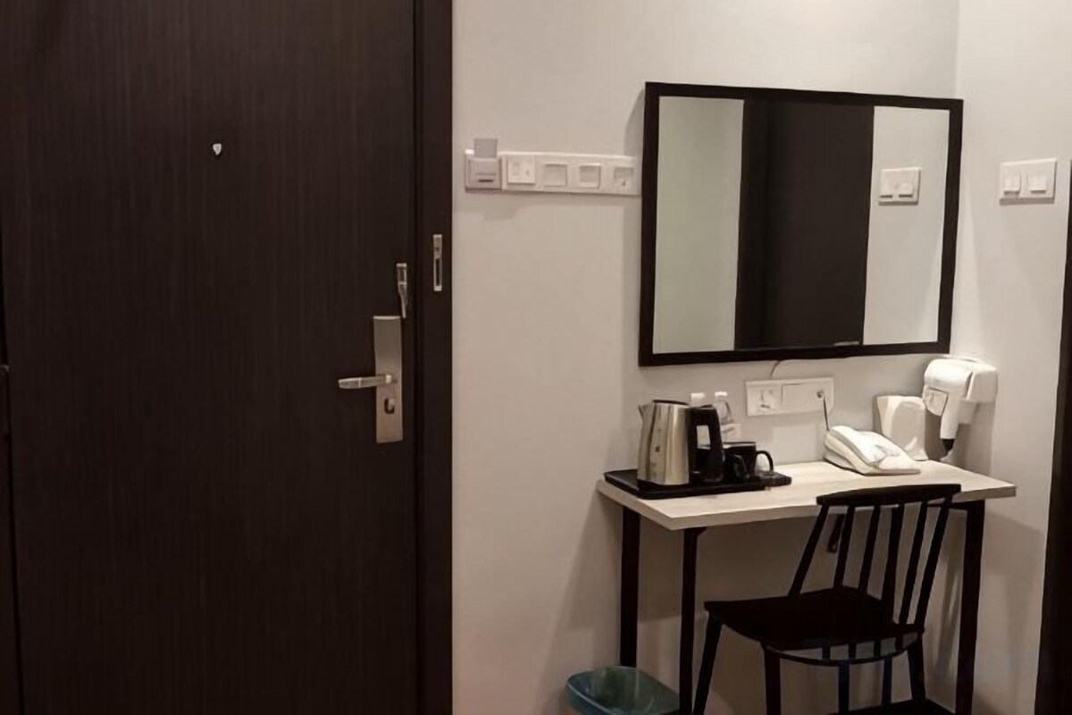 Habitación de hotel con una puerta de madera oscura, un escritorio blanco con un teléfono, una silla negra, un espejo y una tetera negra.