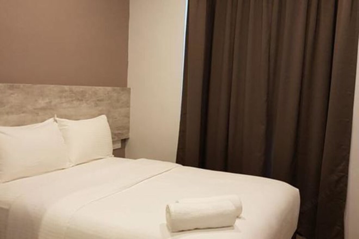 Una cama bien hecha con sábanas blancas y una toalla enrollada al lado, un cabecero y cortinas oscuras.