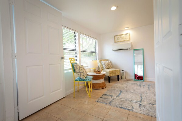 A room with a white door, a chair with a cushion, a small table with a bowl, a rug, and a wall-mounted air conditioner.