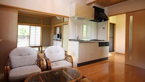 A modern living room with a glass coffee table, two cushioned chairs, a kitchen area with white cabinets, and a wooden ceiling. - Suitaikyou (Nasu)