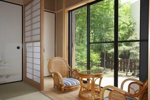 Una habitación japonesa tradicional con suelo de tatami, un fusuma, un asiento de tatami, una mesa de madera baja y un ventanal con vistas a una zona boscosa.