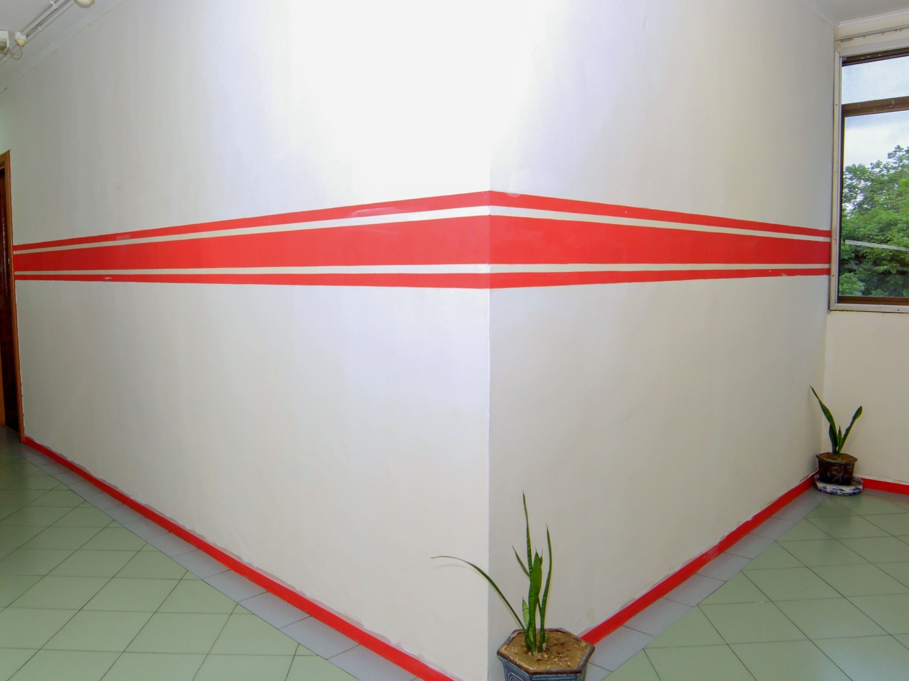Un pasillo con pared blanca y franjas rojas, suelo de baldosas verdes y plantas en macetas.