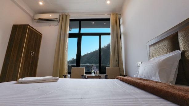 A hotel room with a large bed, a wardrobe, a desk with a chair, and a view of a mountainous landscape through a large window. - Hotel Tapovan Cottage (Narendranagar)