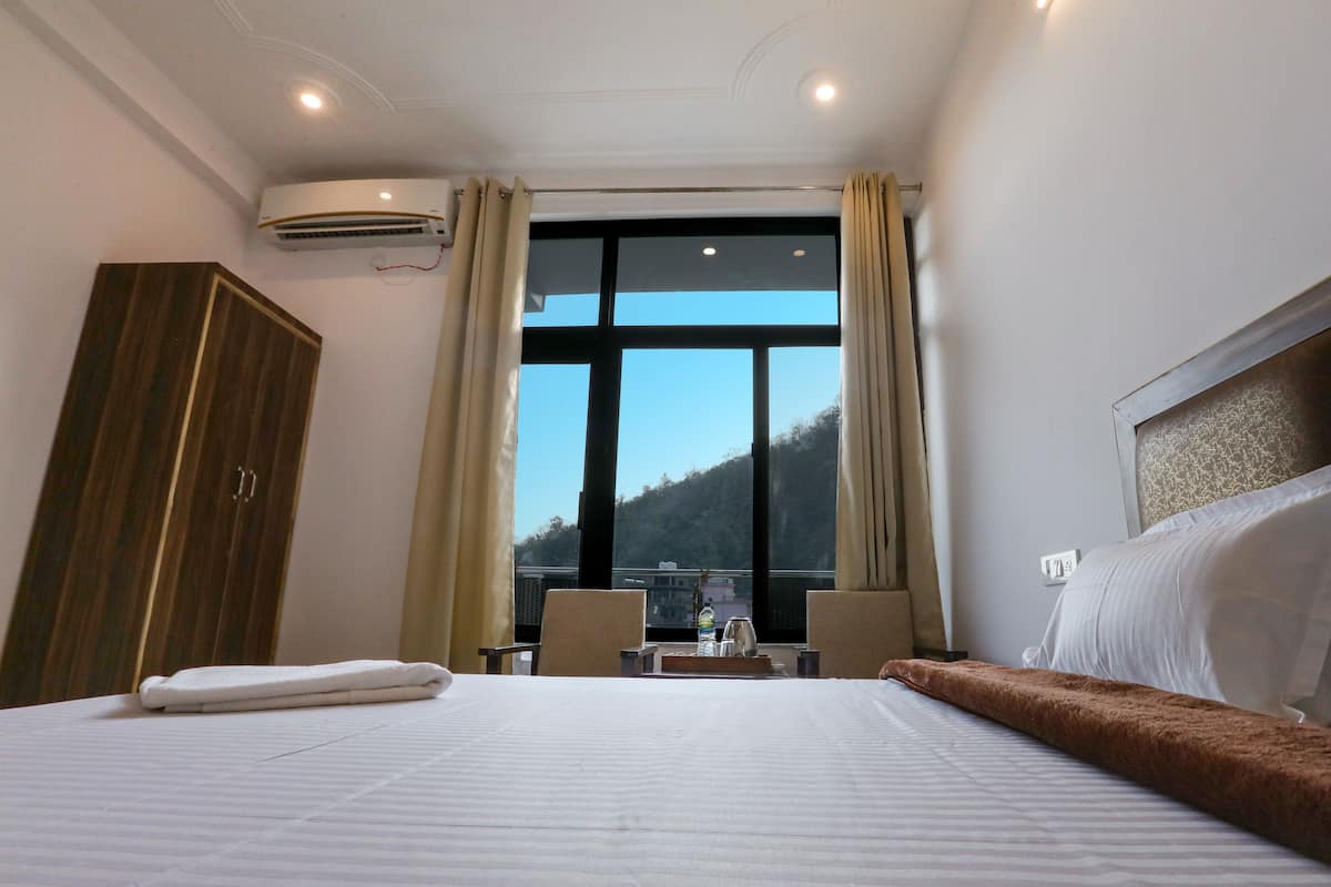 A hotel room with a large bed, a wardrobe, a desk with a chair, and a view of a mountainous landscape through a large window.