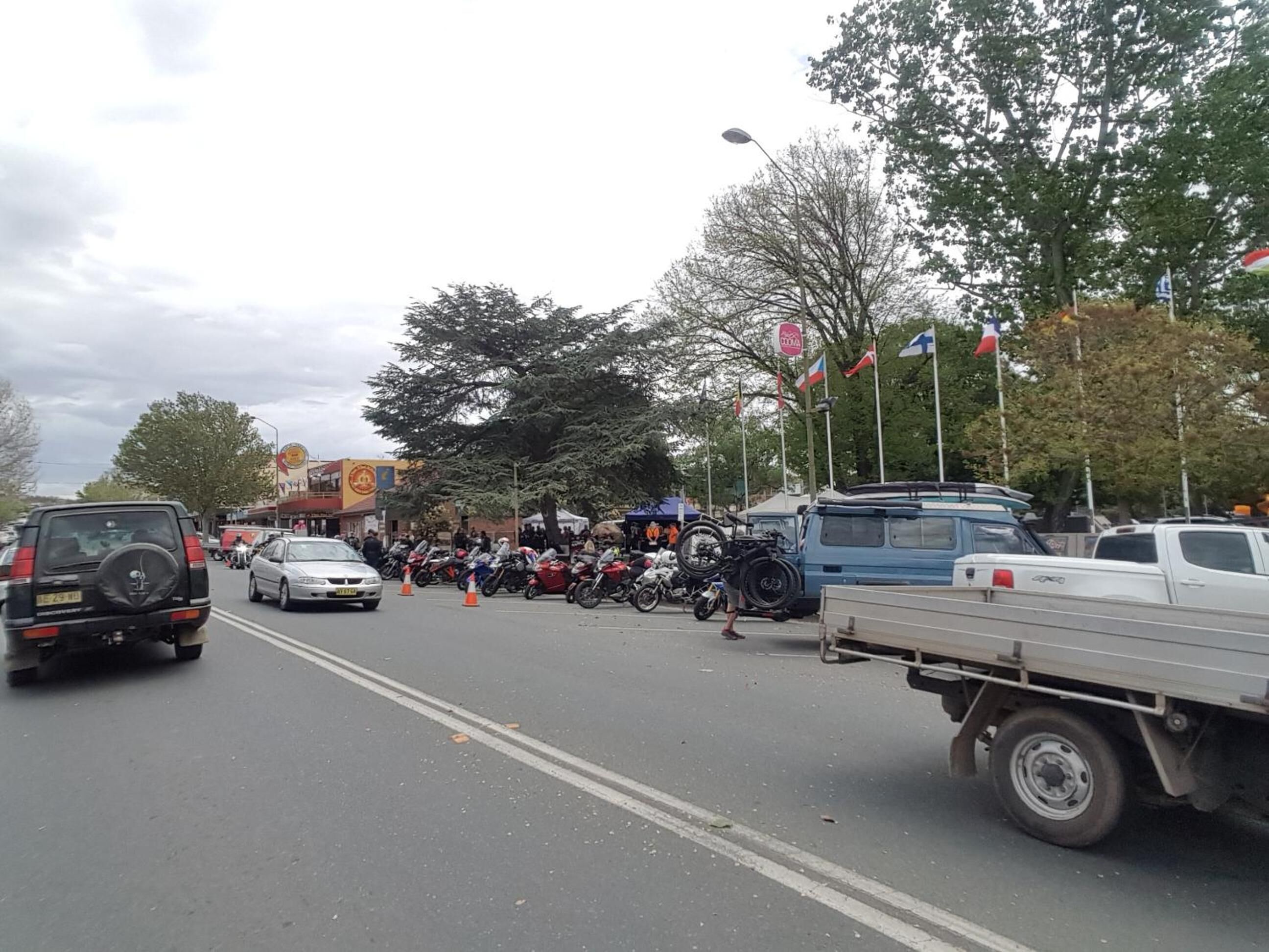 Una strada con motociclette e veicoli parcheggiati, alberi e bandiere.