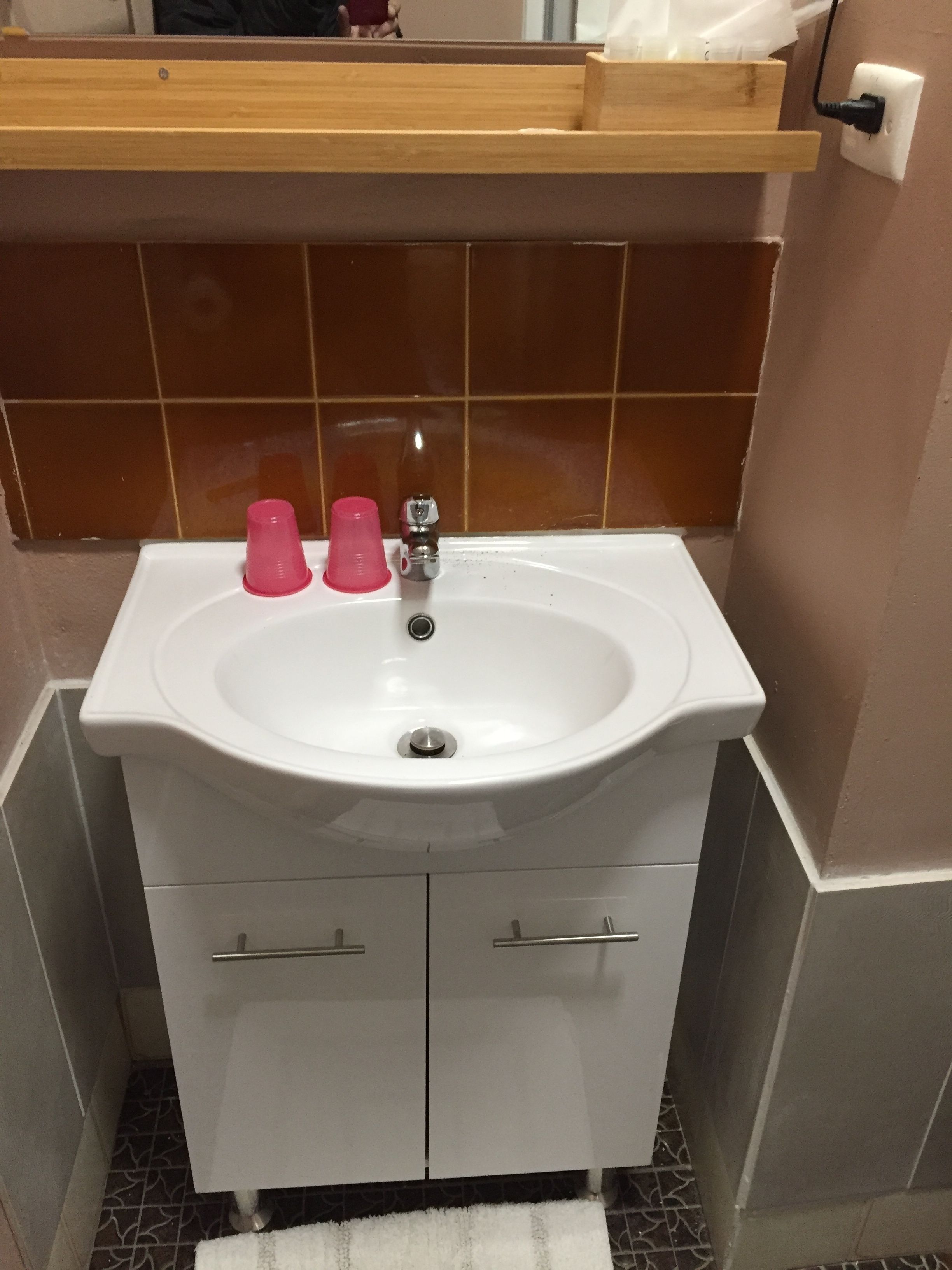 Une salle de bain avec un lavabo blanc, deux tasses roses, un miroir et une étagère en bois au-dessus du lavabo.
