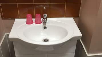 Une salle de bain avec un lavabo blanc, deux tasses roses, un miroir et une étagère en bois au-dessus du lavabo.