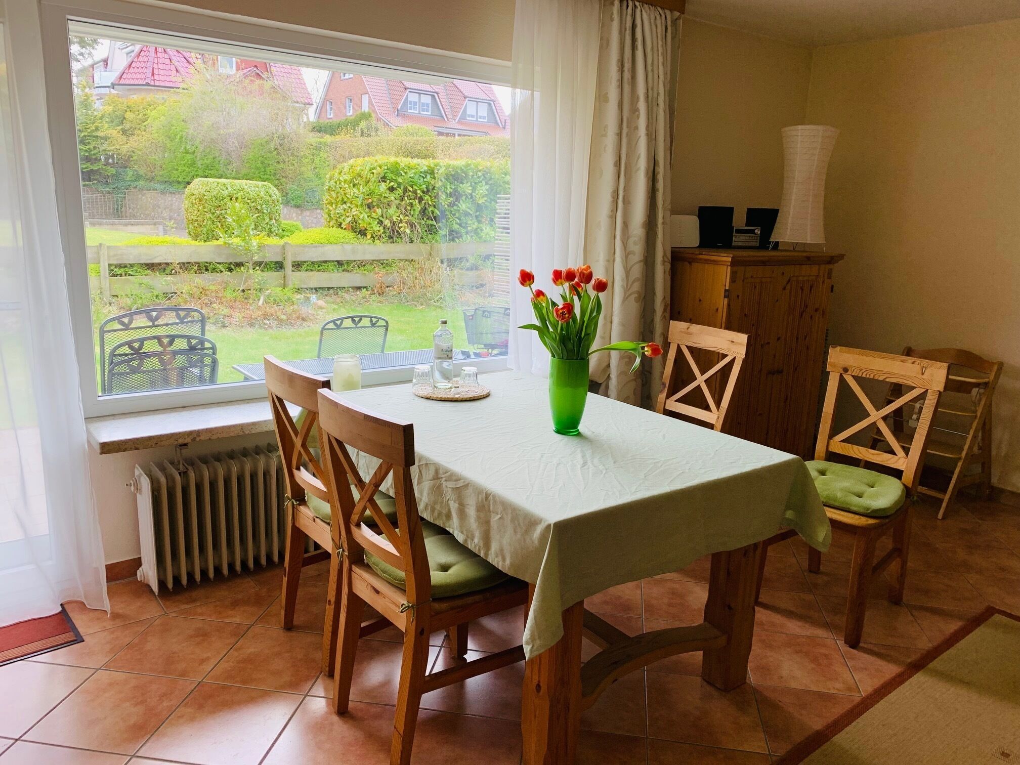 A dining area with a table set for four, a green vase with flowers, and a view of a garden through a window.
