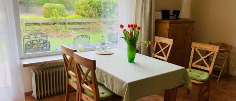 En matplats med ett bord dukat för fyra personer, en grön vas med blommor och utsikt över en trädgård genom ett fönster.